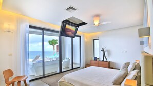 A modern bedroom with a large bed, a TV mounted on the wall, a chair, and a view of the ocean through the sliding doors.