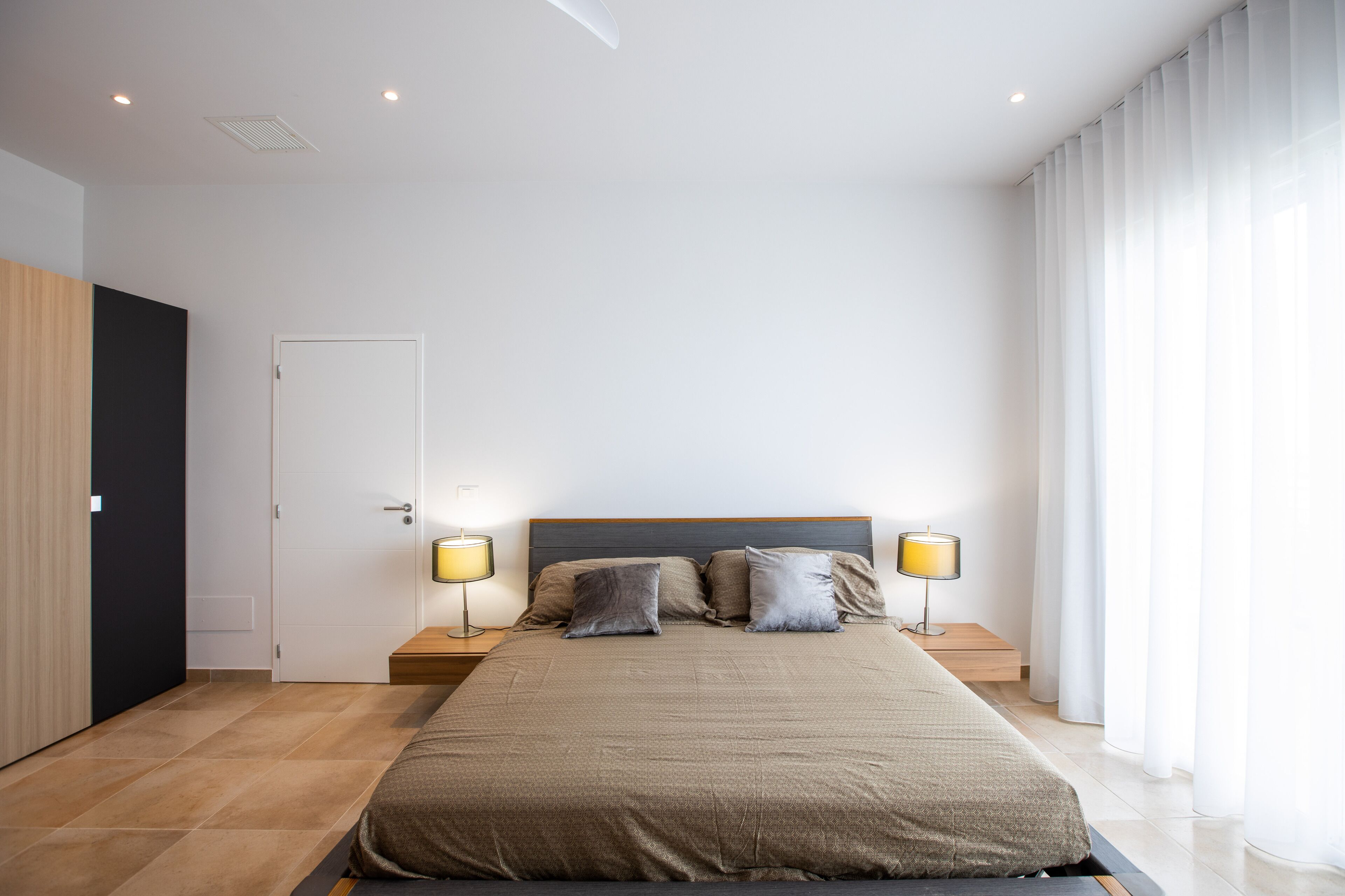 A modern bedroom with a large bed, two bedside tables with lamps, a door, and a window with curtains.