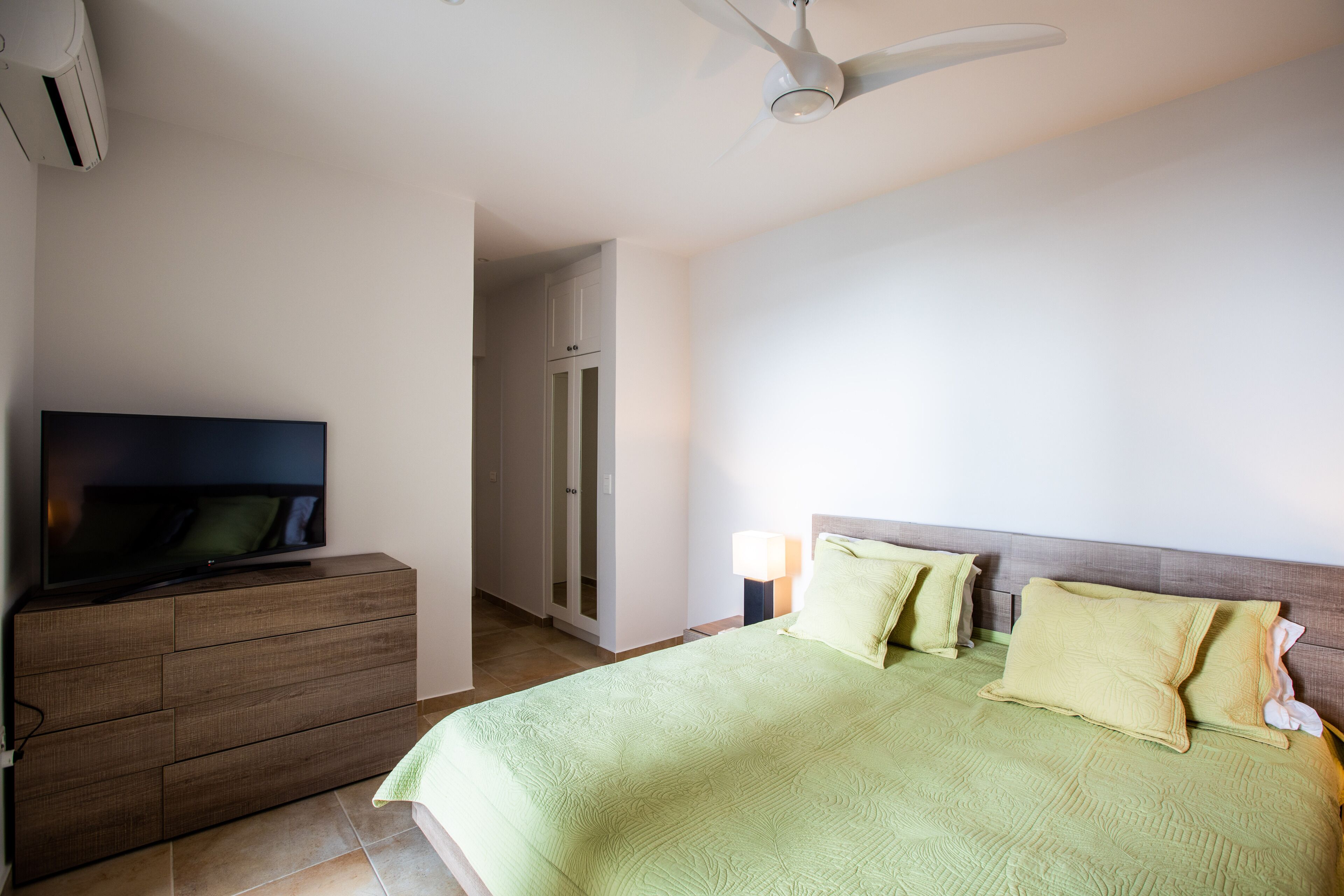 Quarto de hotel com cama, televisão em penteadeira, ventilador de teto e porta para o banheiro.
