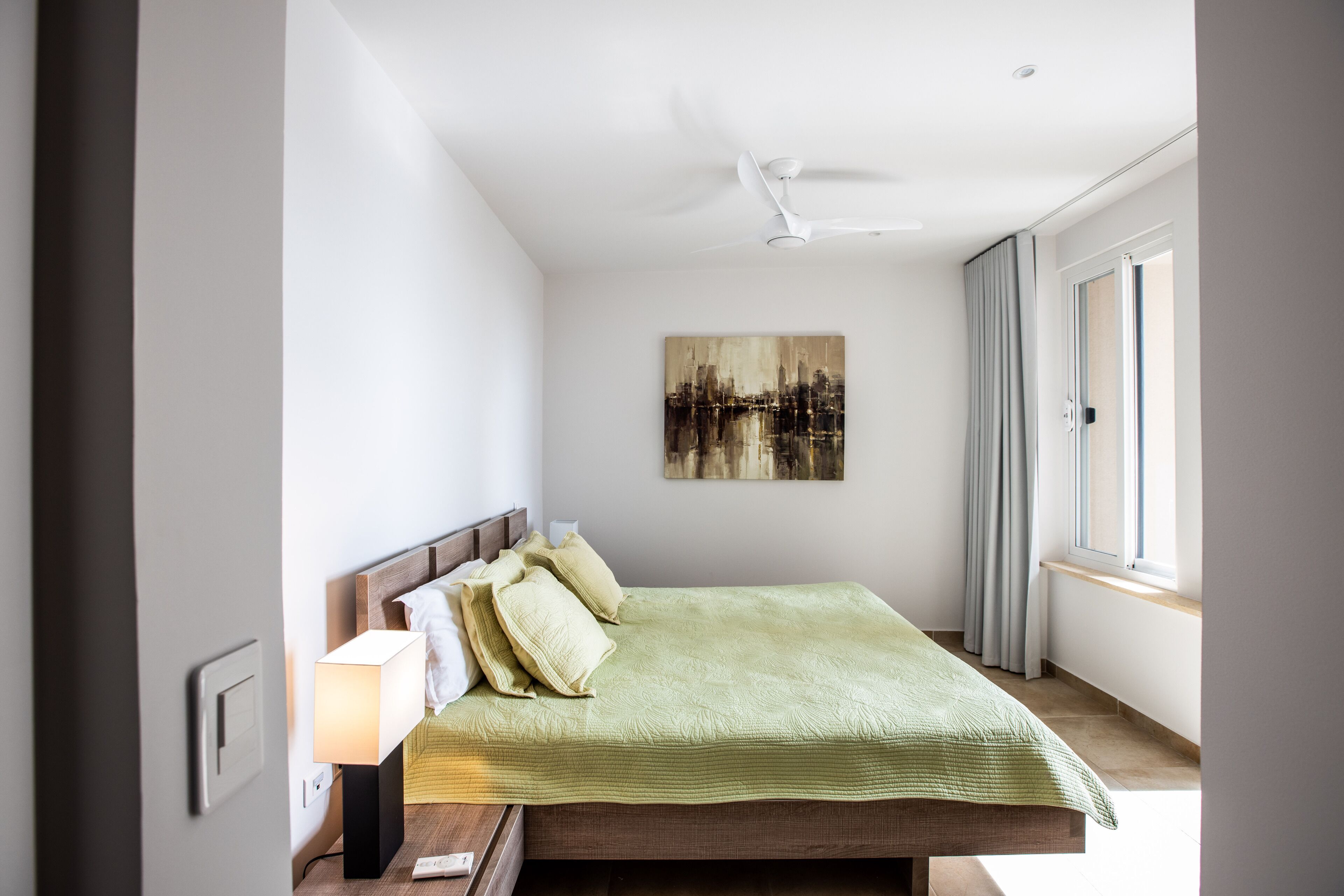Een slaapkamer met een bed, nachtkastje, lamp, plafondventilator, raam en een schilderij aan de muur.