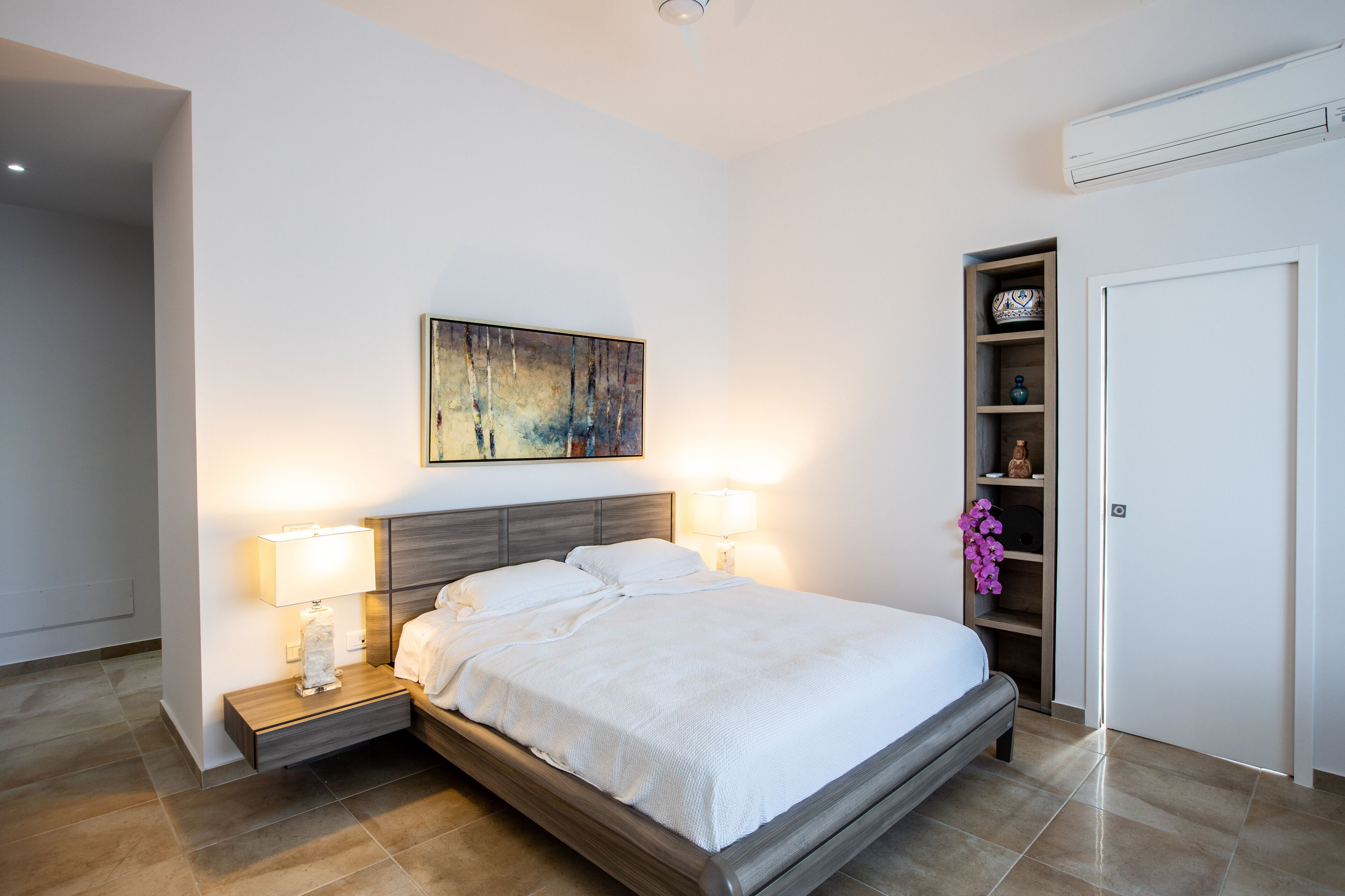 Une chambre à coucher moderne avec un grand lit, des lampes de chevet, une œuvre d’art accrochée au mur, une étagère intégrée ornée d’objets décoratifs et une porte fermée.