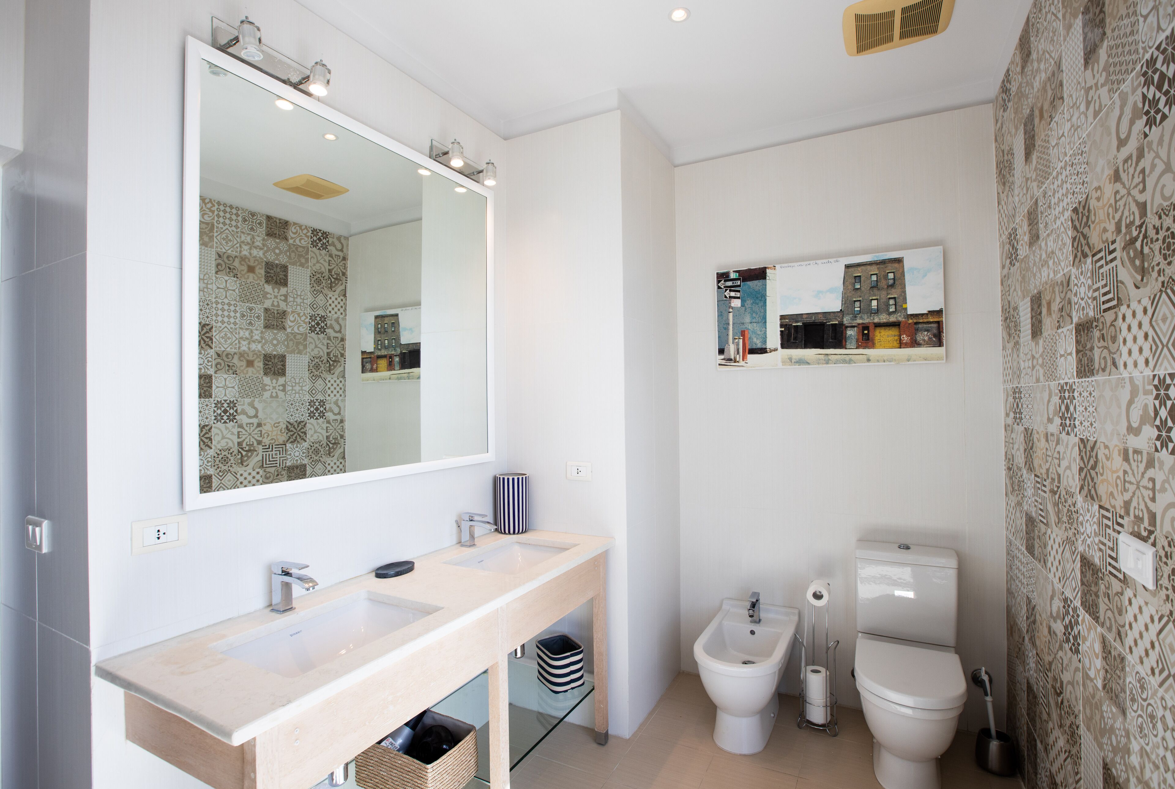 A modern bathroom with a large mirror, two sinks, a toilet, and patterned wall tiles.