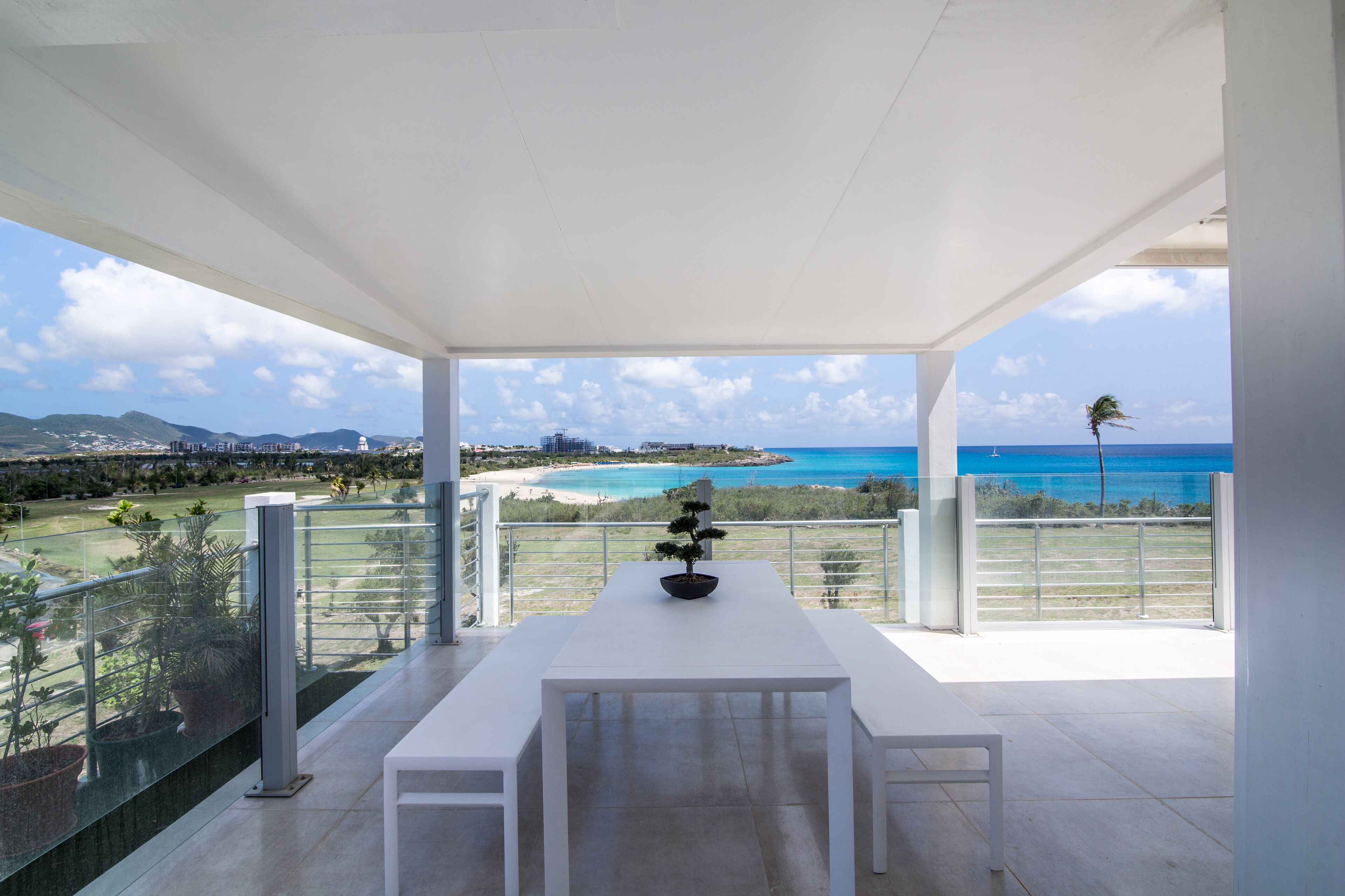 Un balcon avec une table et des chaises blanches, donnant sur une plage et l’océan.