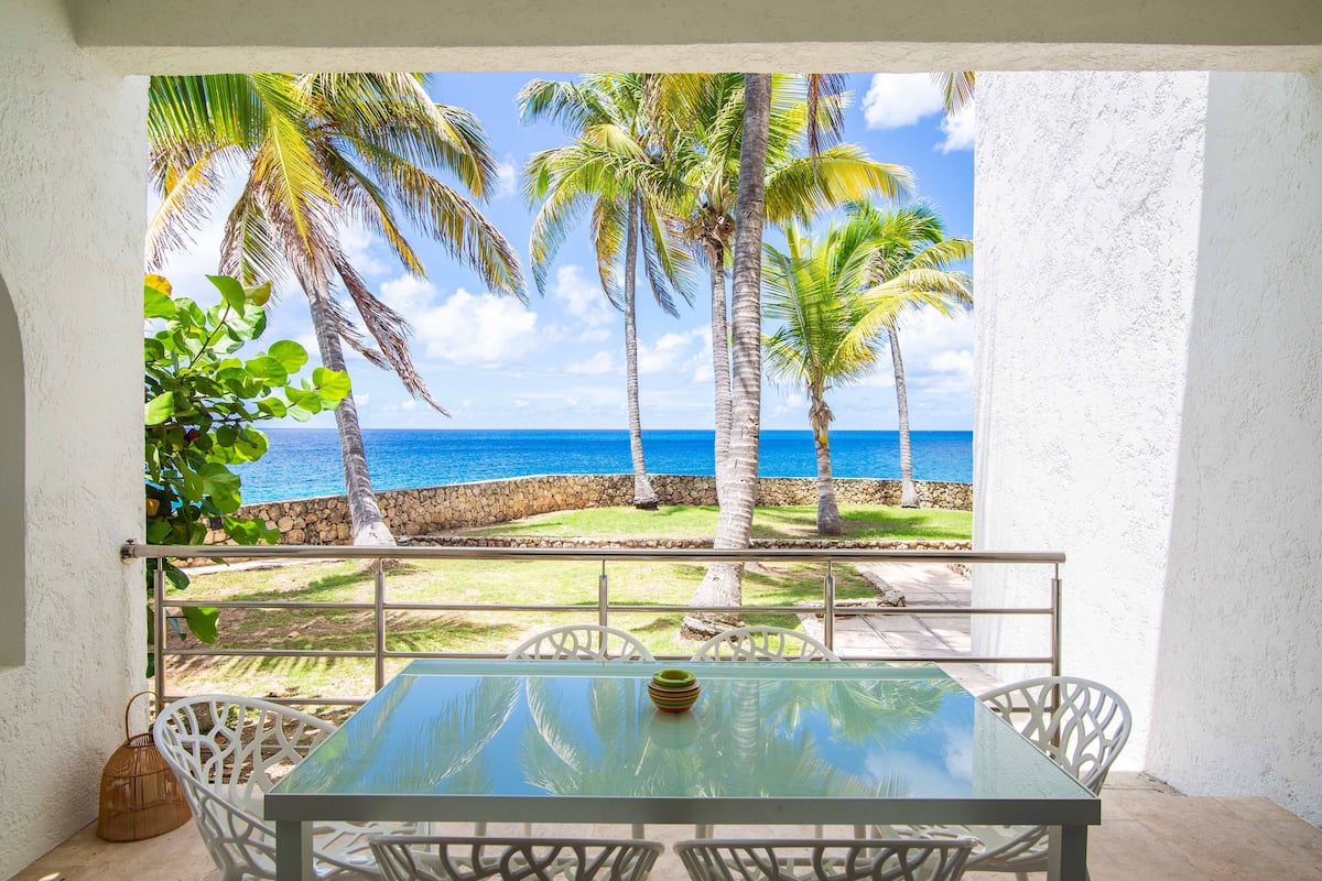 Ein Balkon mit Glastisch und weißen Stühlen, der Blick geht auf einen Strand mit Palmen und das Meer.