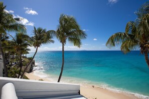 A beach with clear turquoise water, palm trees, and a white building with a balcony overlooking the ocean. - Cupecoy Beach Club (Lowlands)