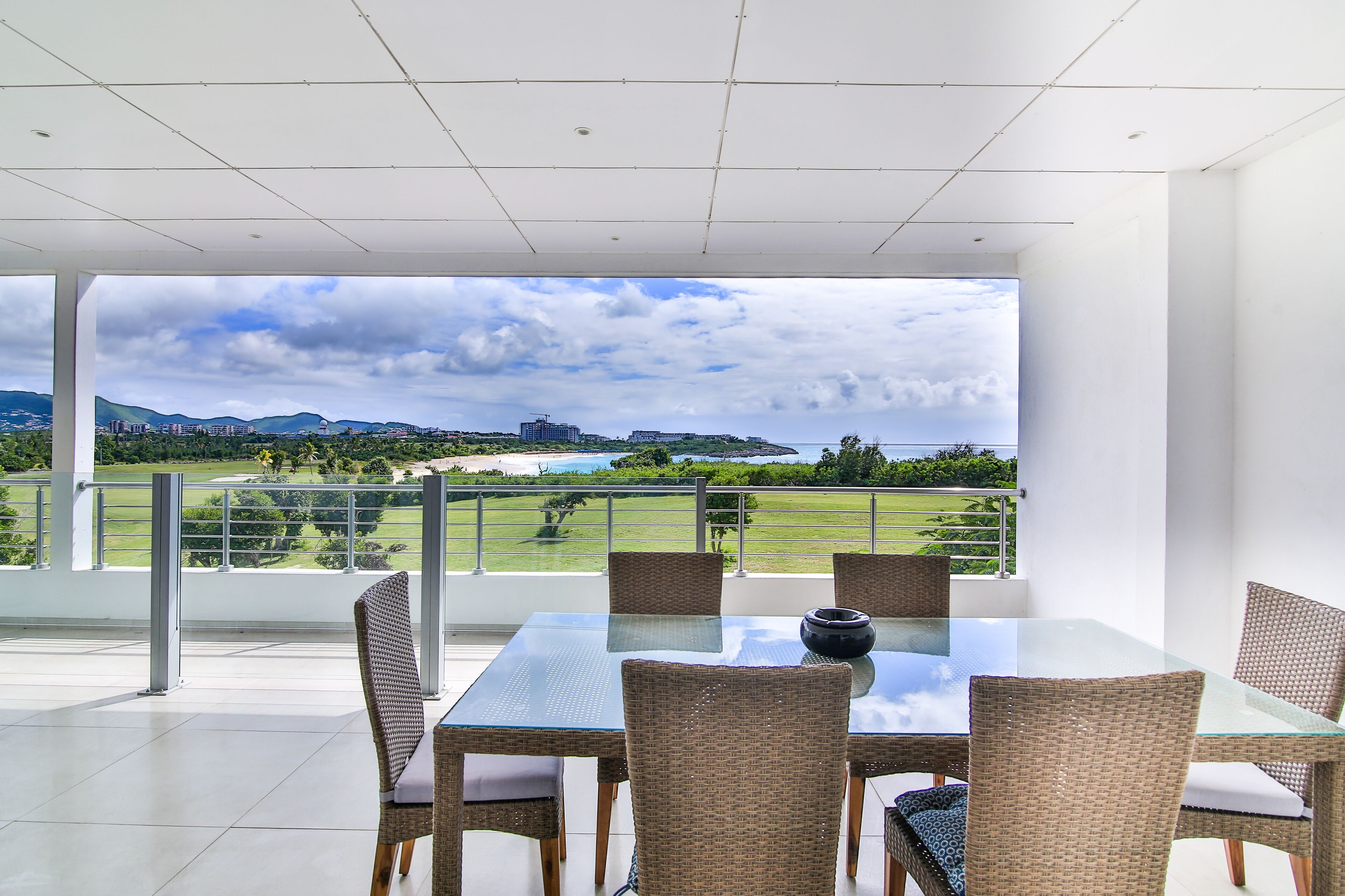Een eetgedeelte met een glazen tafel en rieten stoelen, met uitzicht op een landschap met bergen en een golfbaan.