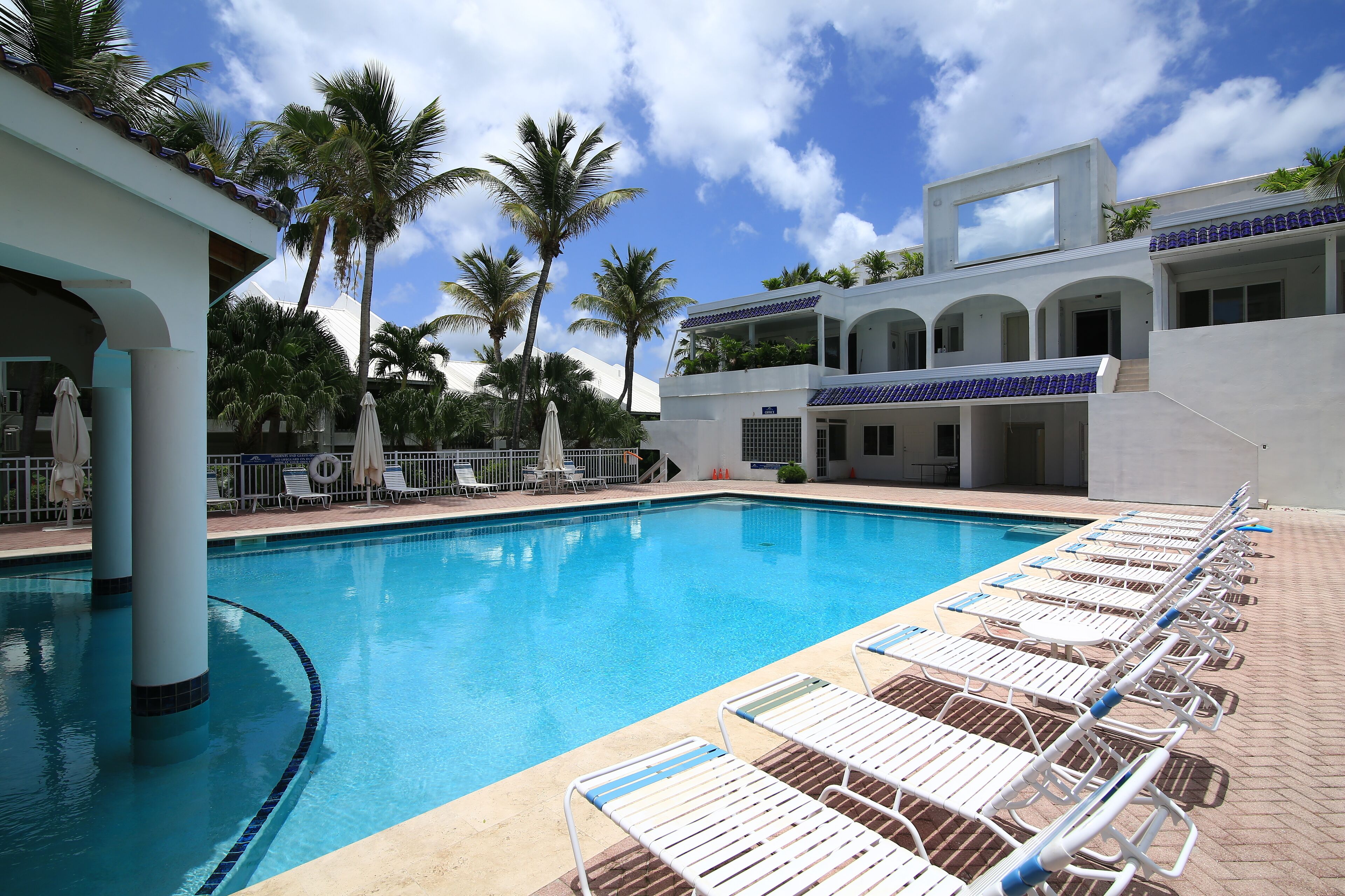 A swimming pool with lounge chairs, palm trees, and a modern building with arched windows.