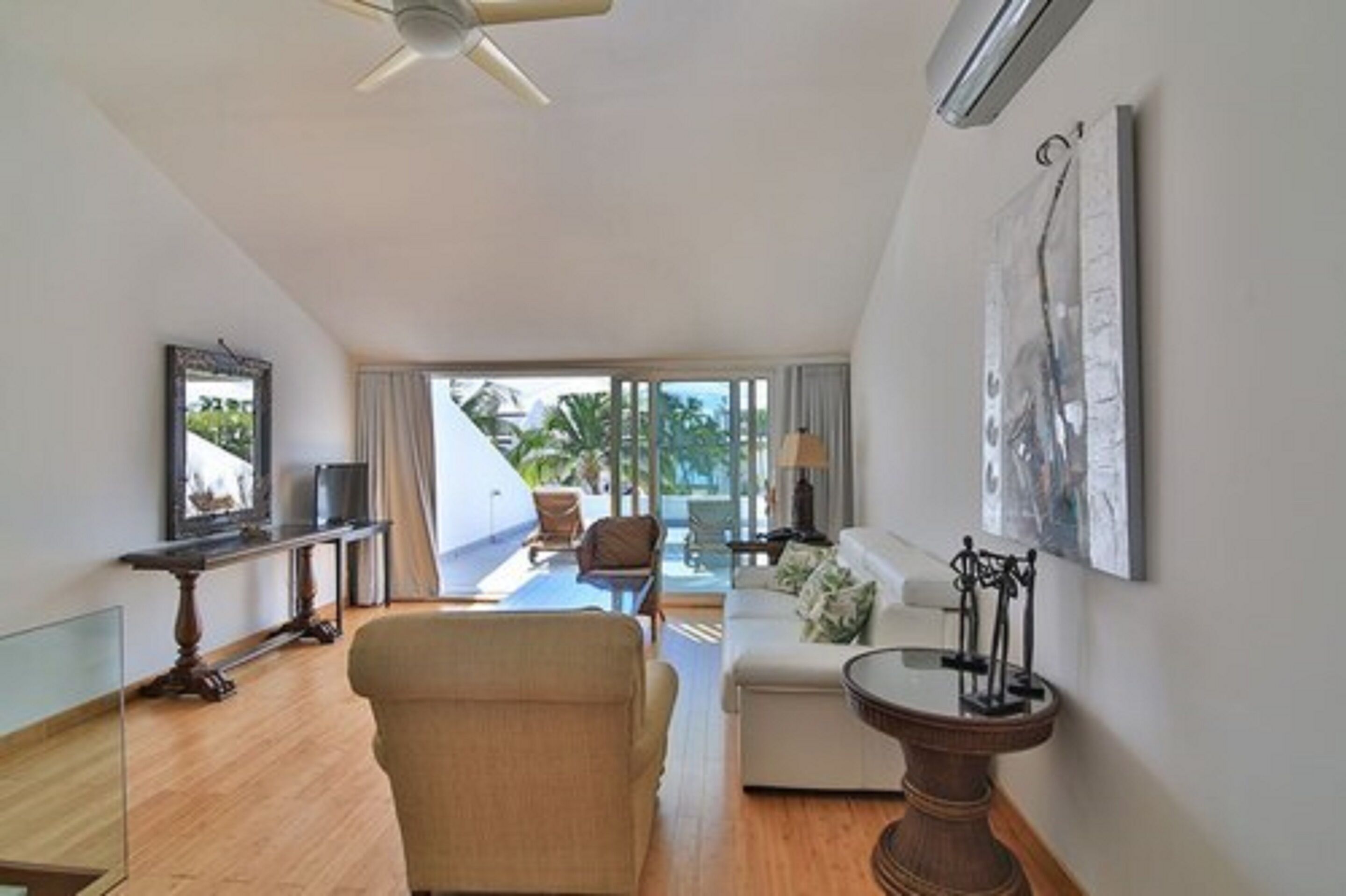 A spacious living area with a sofa, a coffee table, and a TV on a stand. There is a balcony with a view of palm trees.