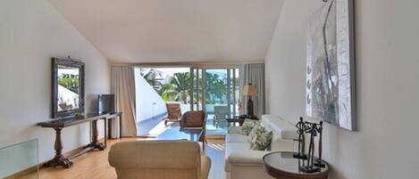 A spacious living area with a sofa, a coffee table, and a TV on a stand. There is a balcony with a view of palm trees.