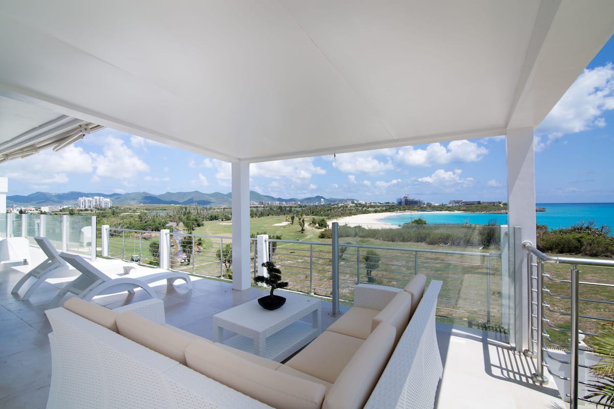 Ein Balkon mit weißen Korbmöbeln, einem kleinen Tisch mit Bonsai und Blick auf Strand und Berge.