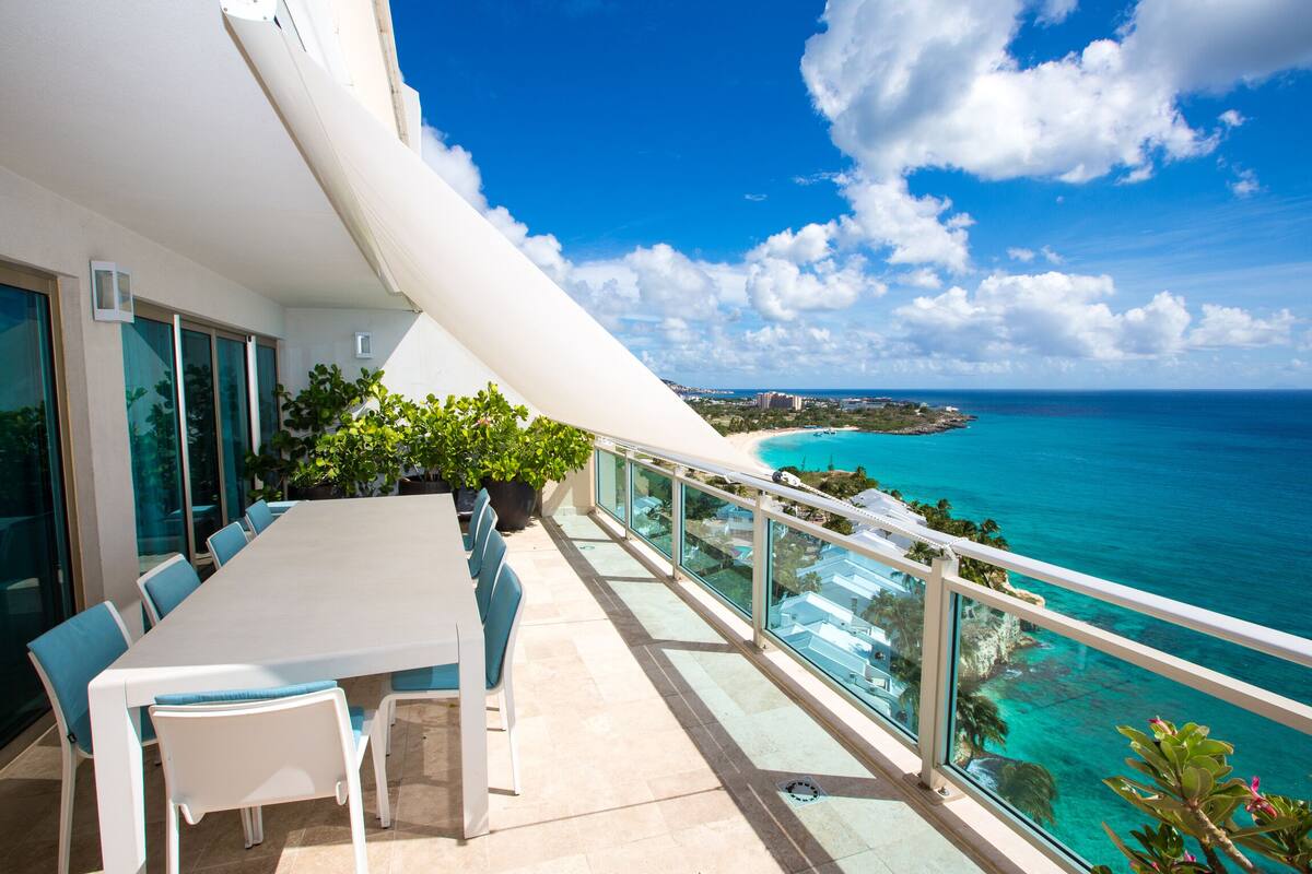 Un balcon doté d’une table et de chaises, offrant une vue sur la côte, avec une mer d’un bleu limpide et une île visible au loin.