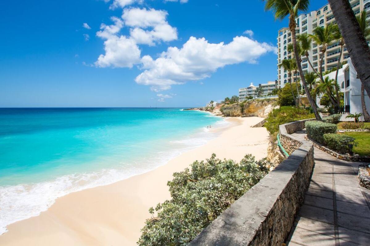 Une plage bordée d’un ciel bleu azur, d’une eau turquoise et d’un sentier de pierres menant à un bâtiment entouré de palmiers.