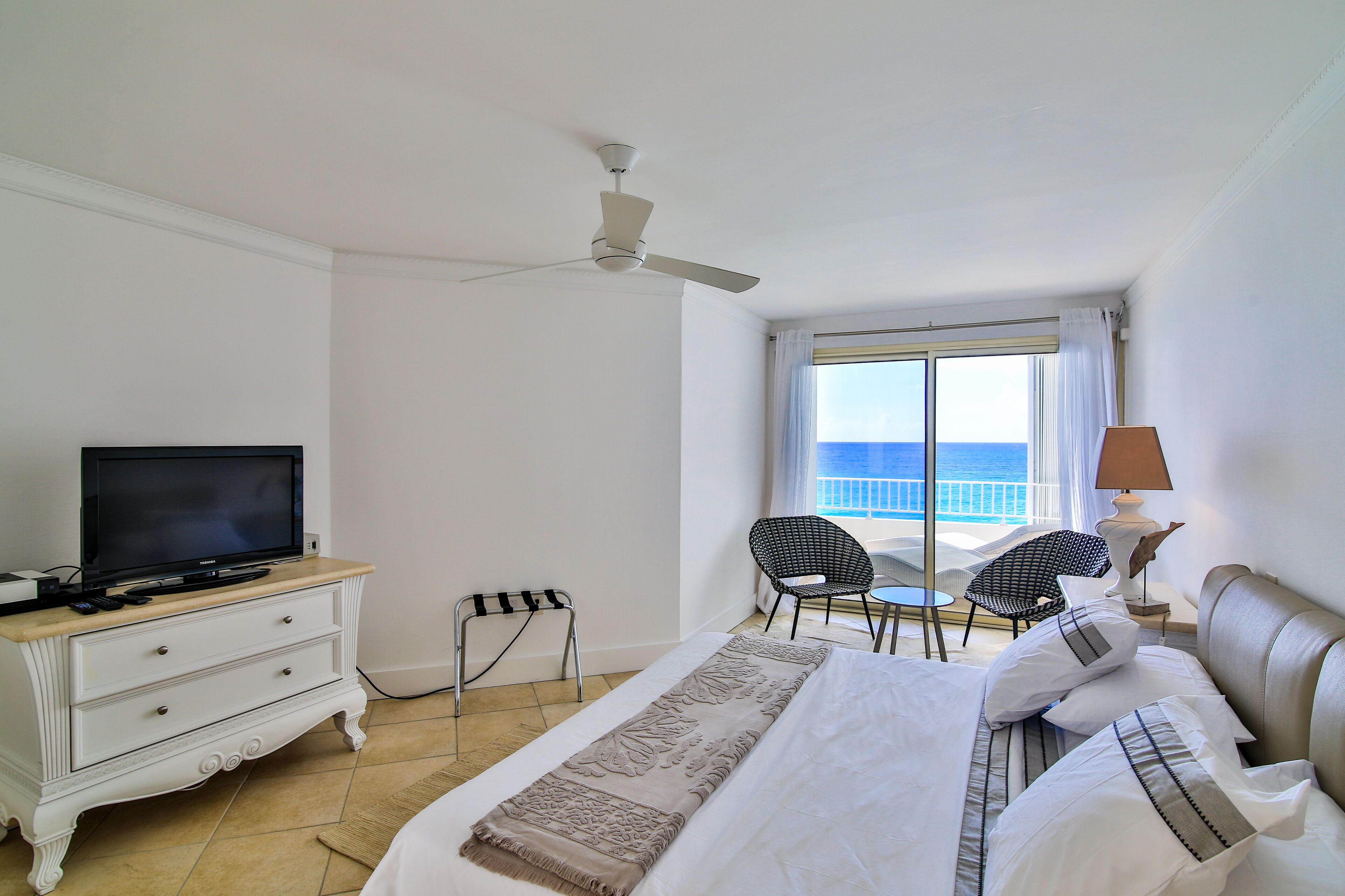 A hotel room with a bed, a television, a ceiling fan, a window with a view of the sea, and a small table with chairs.