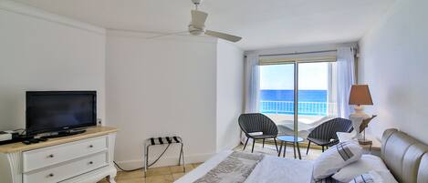 A hotel room with a bed, a television, a ceiling fan, a window with a view of the sea, and a small table with chairs.