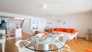 A modern living room with a glass dining table, white chairs, a sofa with orange cushions, and a ceiling fan. - Sapphire Fatastic (Lowlands)