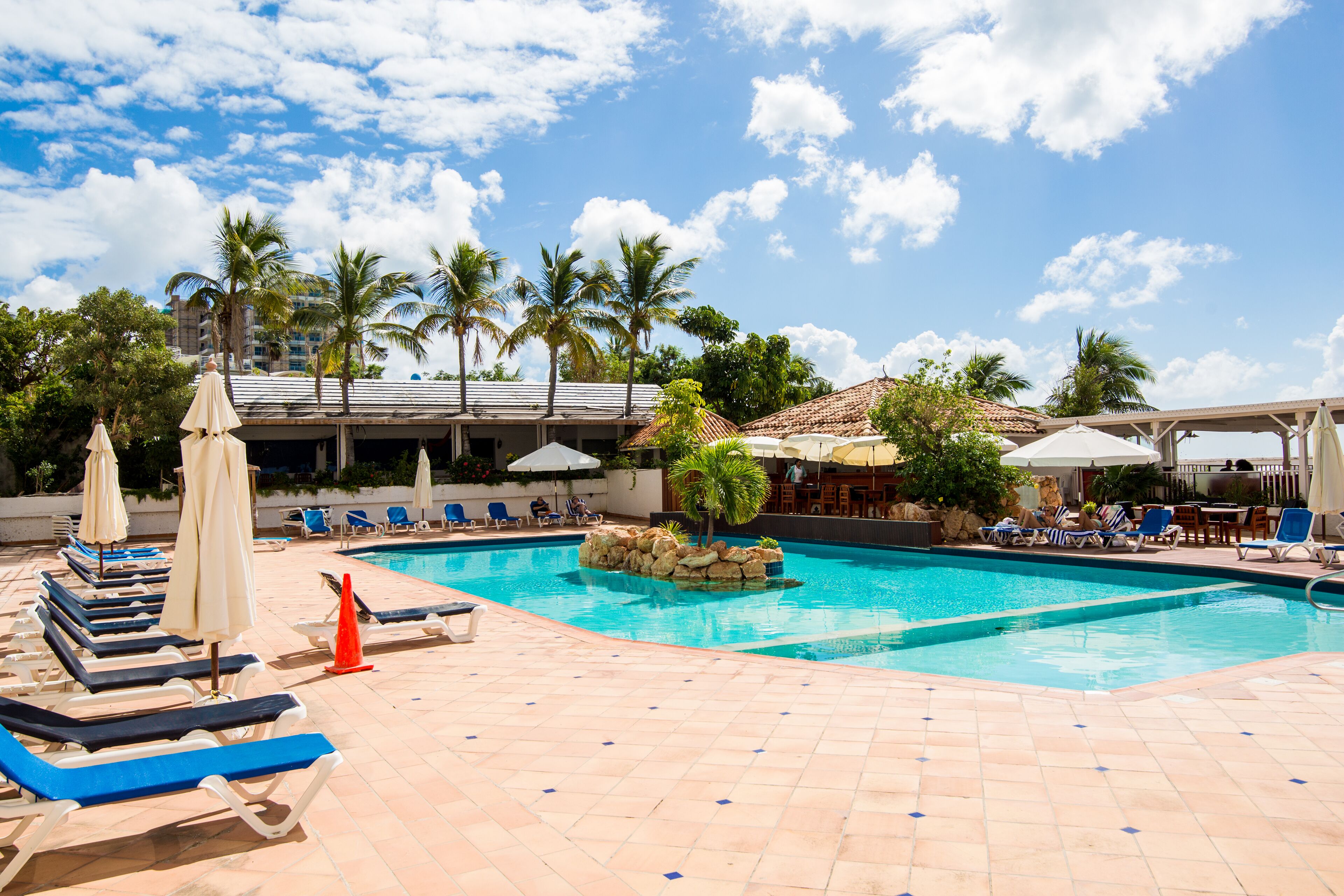 A pool area with lounge chairs and umbrellas, palm trees, and a building in the background.