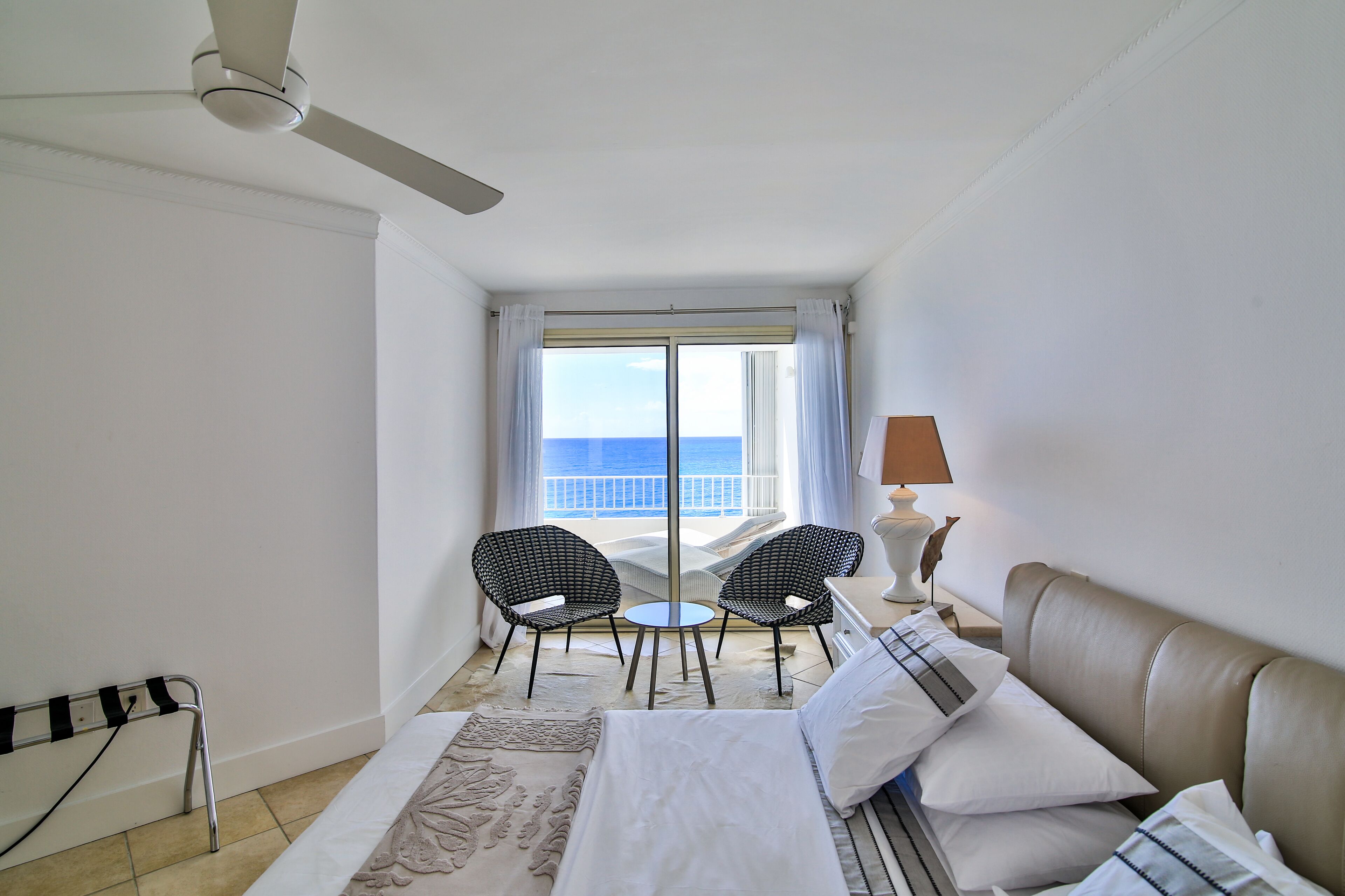 Une chambre d’hôtel avec un lit, deux chaises, une table, une lampe et une vue sur la mer.