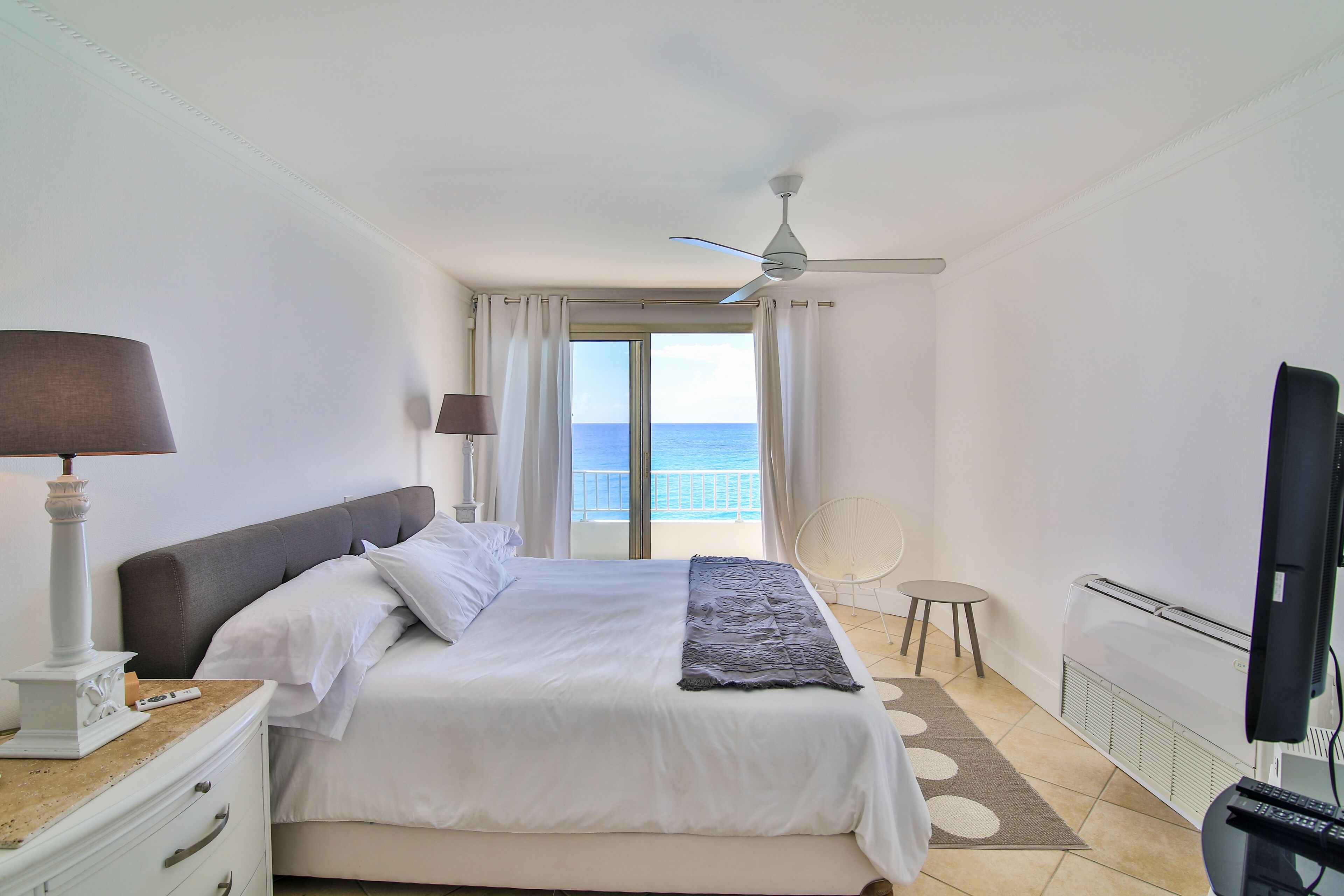 A hotel room with a bed, a ceiling fan, a television, and a view of the sea through the window.