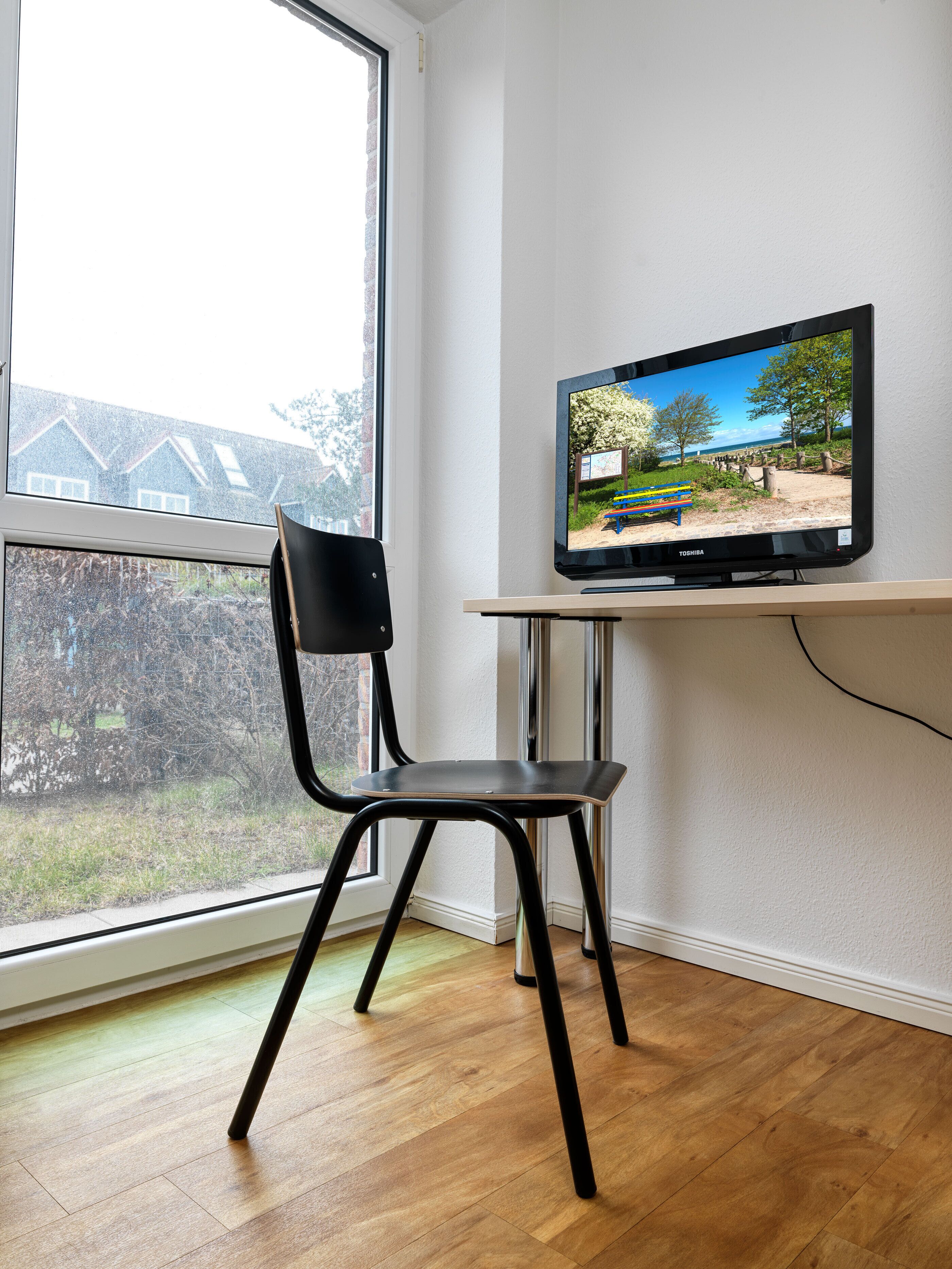 A room with a television on a stand, a chair, and a window overlooking a garden.