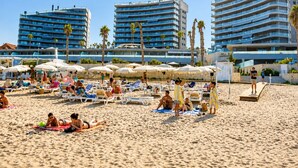 Une plage avec des transats et des parasols, des palmiers et des bâtiments modernes en arrière-plan.