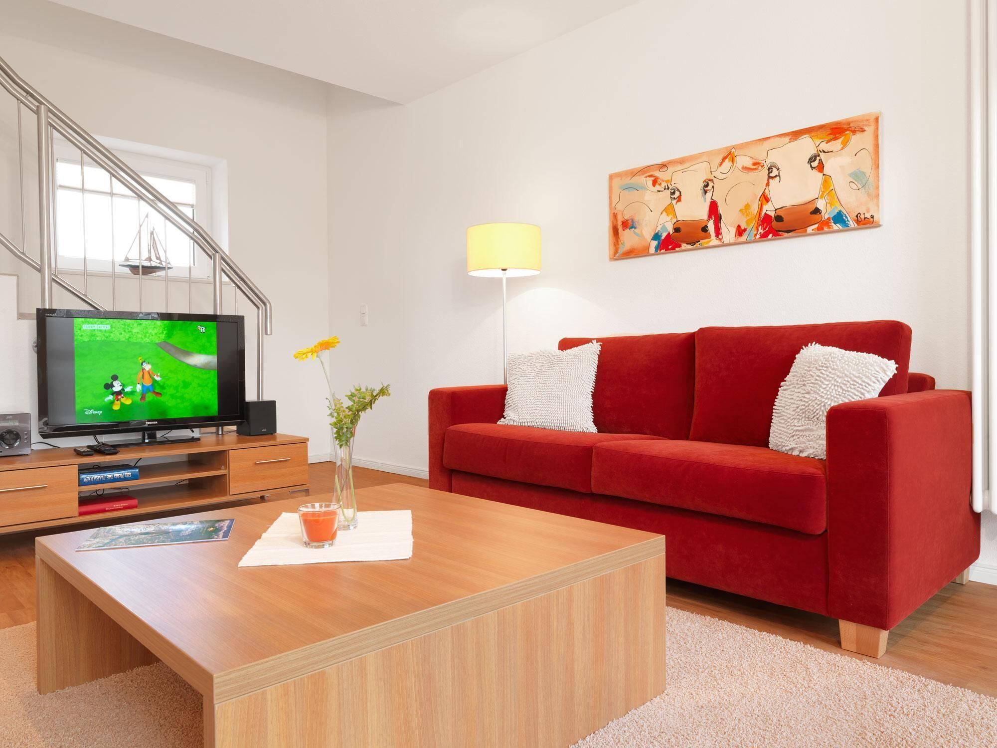 A modern living room with a red sofa, a wooden coffee table, a TV on a stand, a painting on the wall, and a lamp.