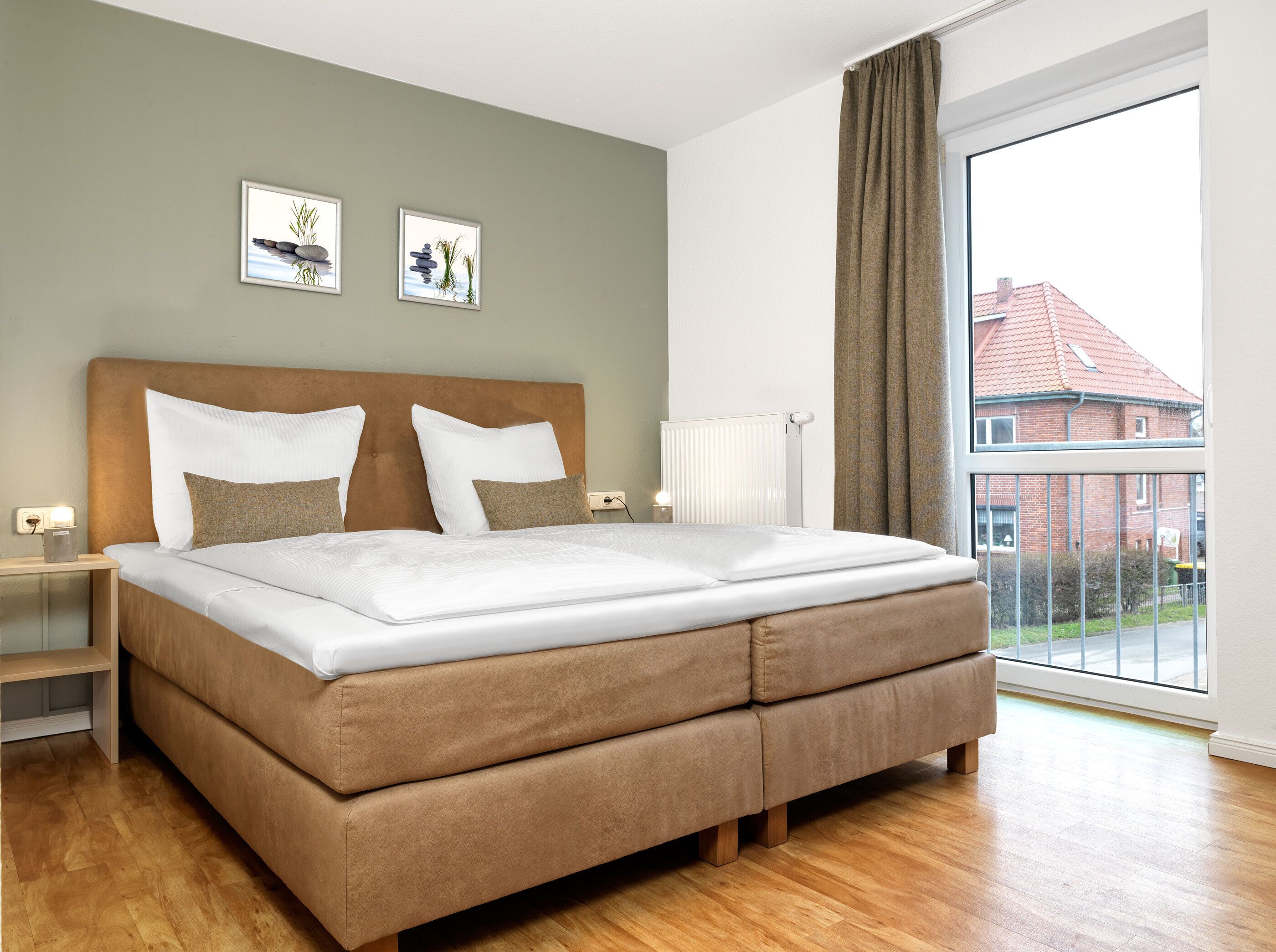 Une chambre avec un grand lit, une table de chevet, une commode, un radiateur et une fenêtre donnant sur des immeubles.
