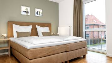 Une chambre avec un grand lit, une table de chevet, une commode, un radiateur et une fenêtre donnant sur des immeubles.