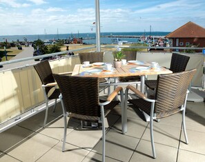 Terrassa amb cadires de vimet i una taula preparada per dinar, amb vista a una marina amb vaixells i una costa llunyana.