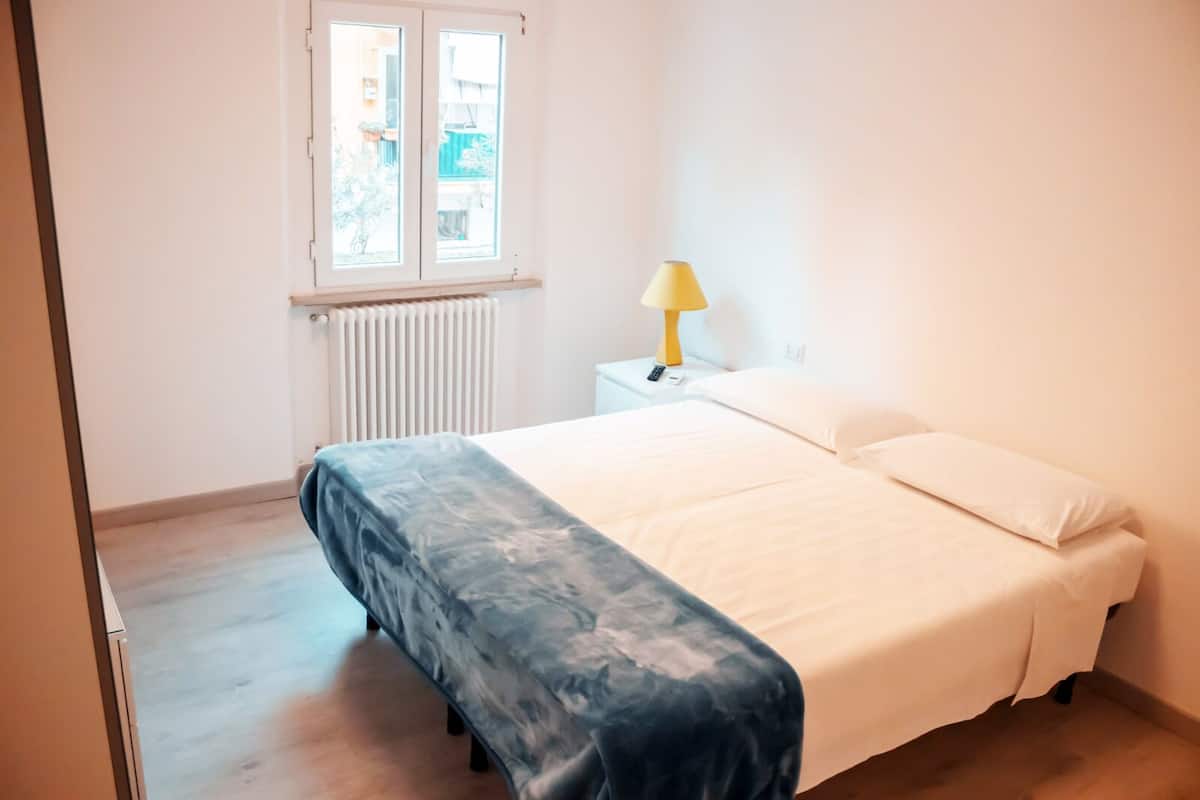 Um quarto com uma cama, mesa de cabeceira com abajur, janela e radiador.