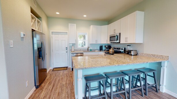 A kitchen with white cabinets, a granite countertop, a microwave, a stove, and a sink. There are three bar stools at the island.