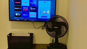 A TV mounted on a wall displaying various streaming services, a black fan, a green chair, and a black desk with a basket. - Pousada EAS GRU Airport (Guarulhos)
