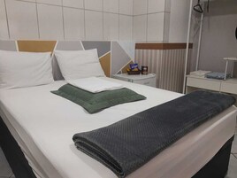 Una cama bien tendida con almohadas blancas y verdes, una manta doblada y una mesita de noche con una lámpara y una caja pequeña.