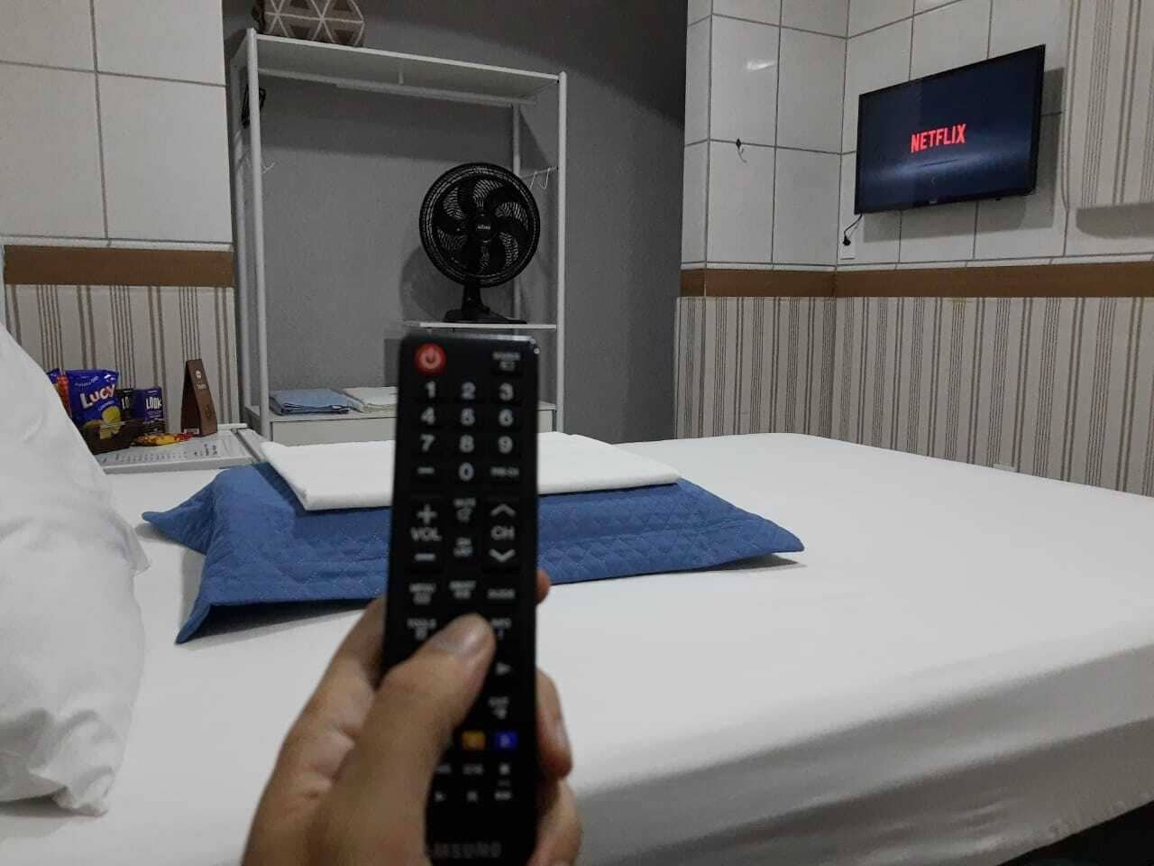 A hotel room with a bed, a remote control, a fan, and a TV displaying 'NETFLIX'.