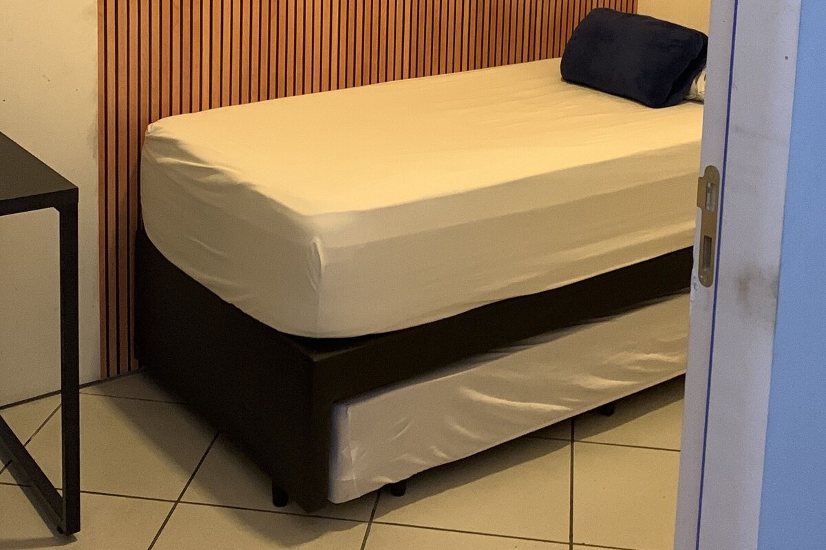 Een kleine kamer met een enkel bed, een houten lambrisering, een nachtkastje en een deur.