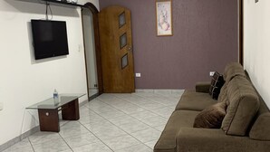 A living room with a glass-top coffee table, a brown sofa, a flat-screen TV mounted on the wall, and a wooden door. - Pousada EAS GRU Airport (Guarulhos)