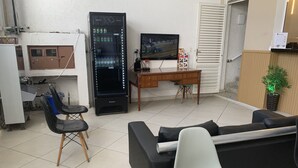A living room with a sofa, a TV, a vending machine, a desk, and a chair. - Pousada EAS GRU Airport (Guarulhos)