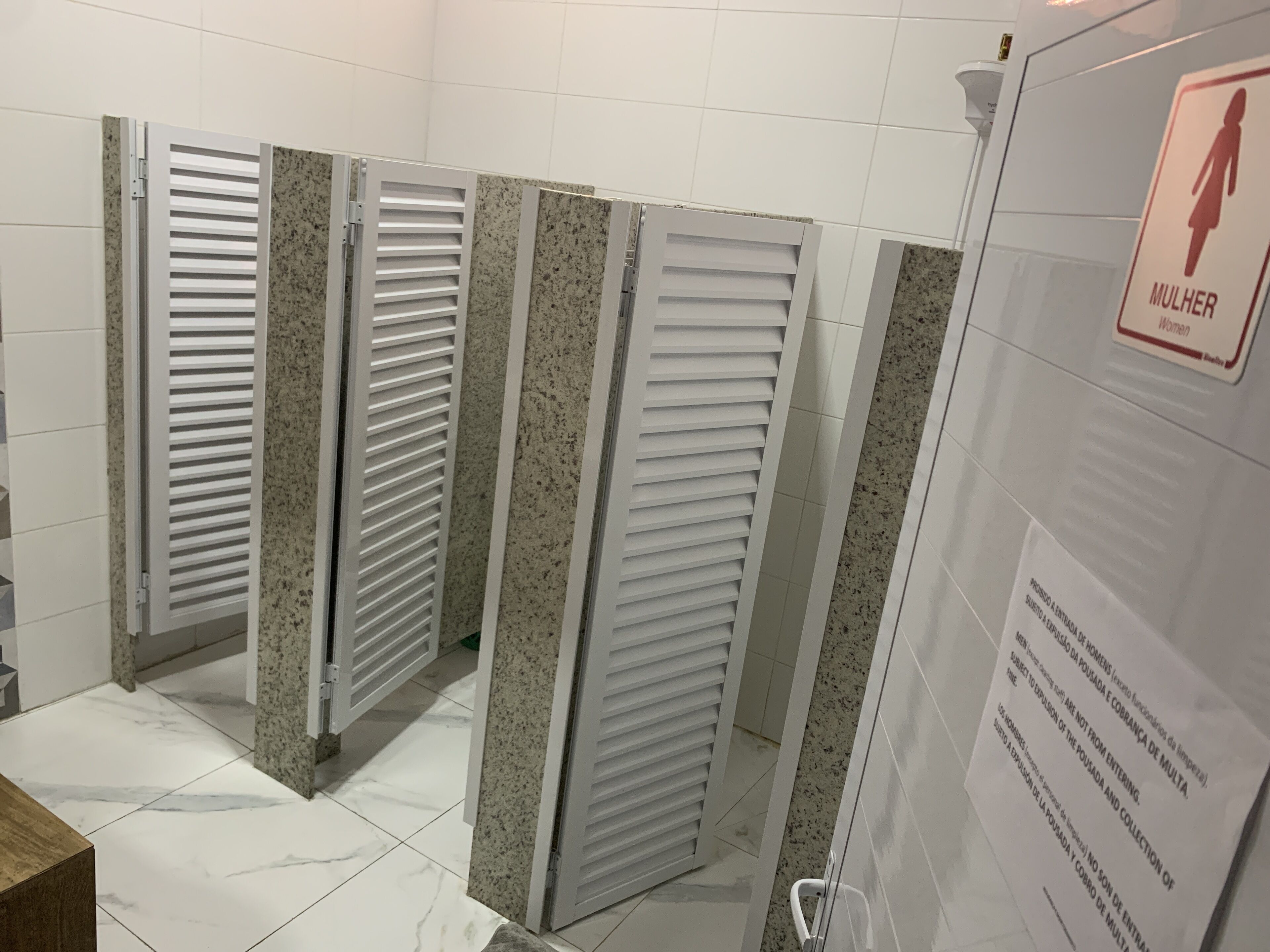 Baño con tres cabinas, cada una con puerta blanca y panel de vidrio esmerilado. Un letrero indica la sección de mujeres.