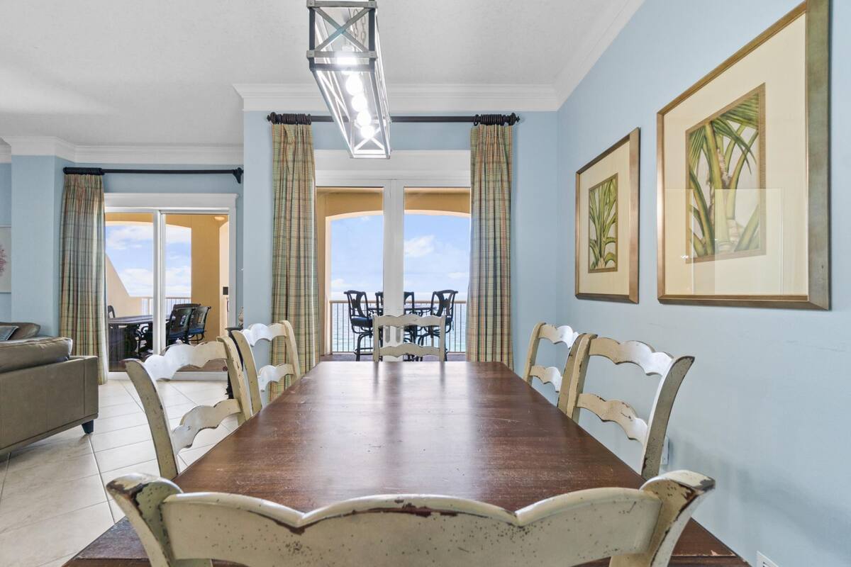 A dining area with a long wooden table, white chairs, and a view through large windows.