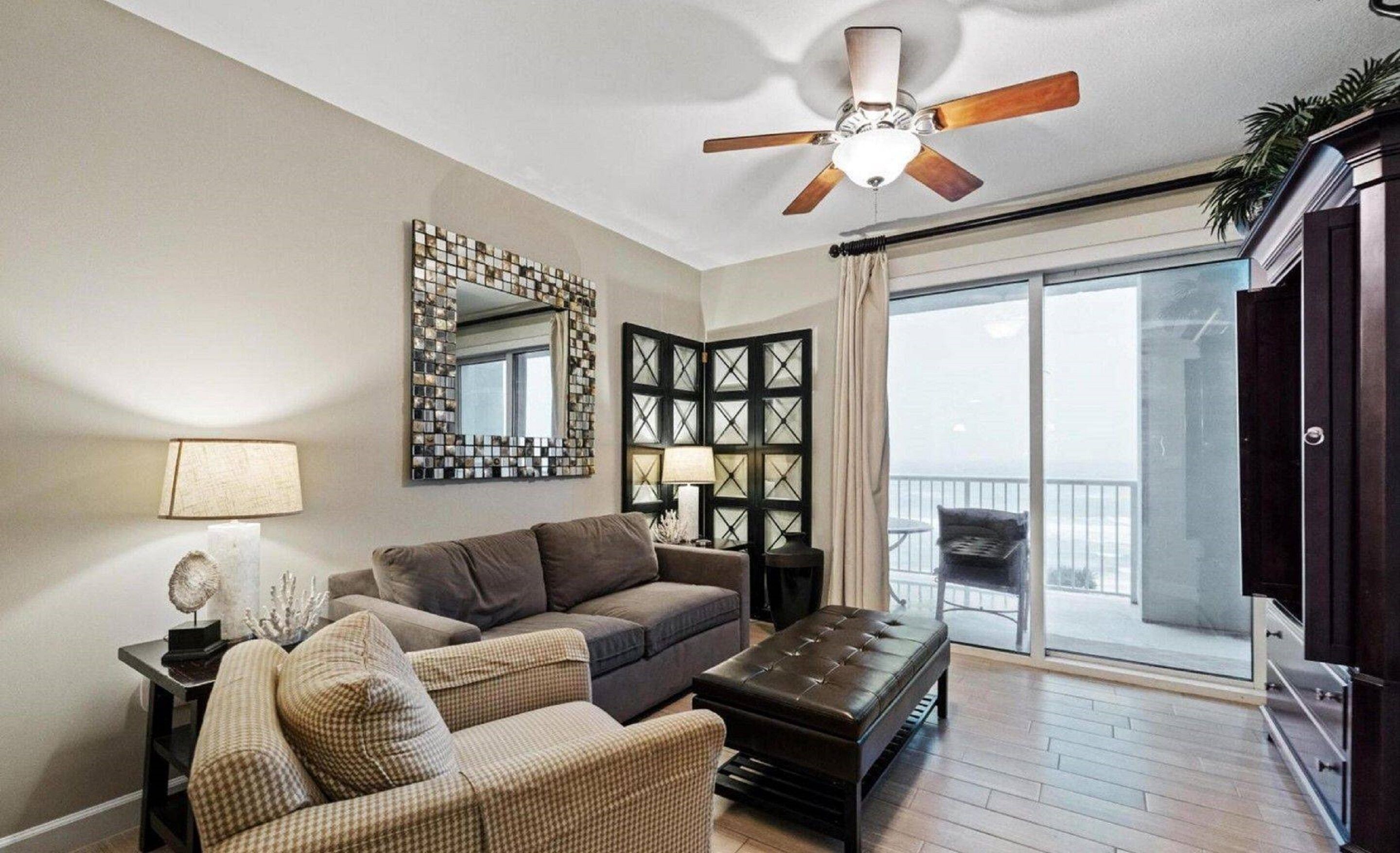 Una sala de estar con ventilador de techo, un espejo grande, un sofá, una silla, una mesa de centro y un balcón con una silla.