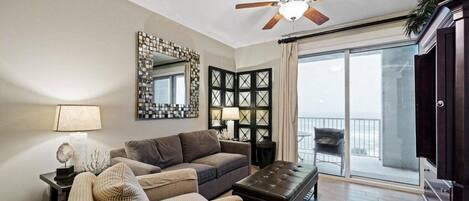 Una sala de estar con ventilador de techo, un espejo grande, un sofá, una silla, una mesa de centro y un balcón con una silla.