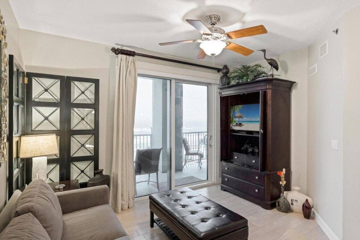 A living room with a sofa, a coffee table, a dark wooden cabinet with a painting, a ceiling fan, and a sliding glass door leading to a balcony.