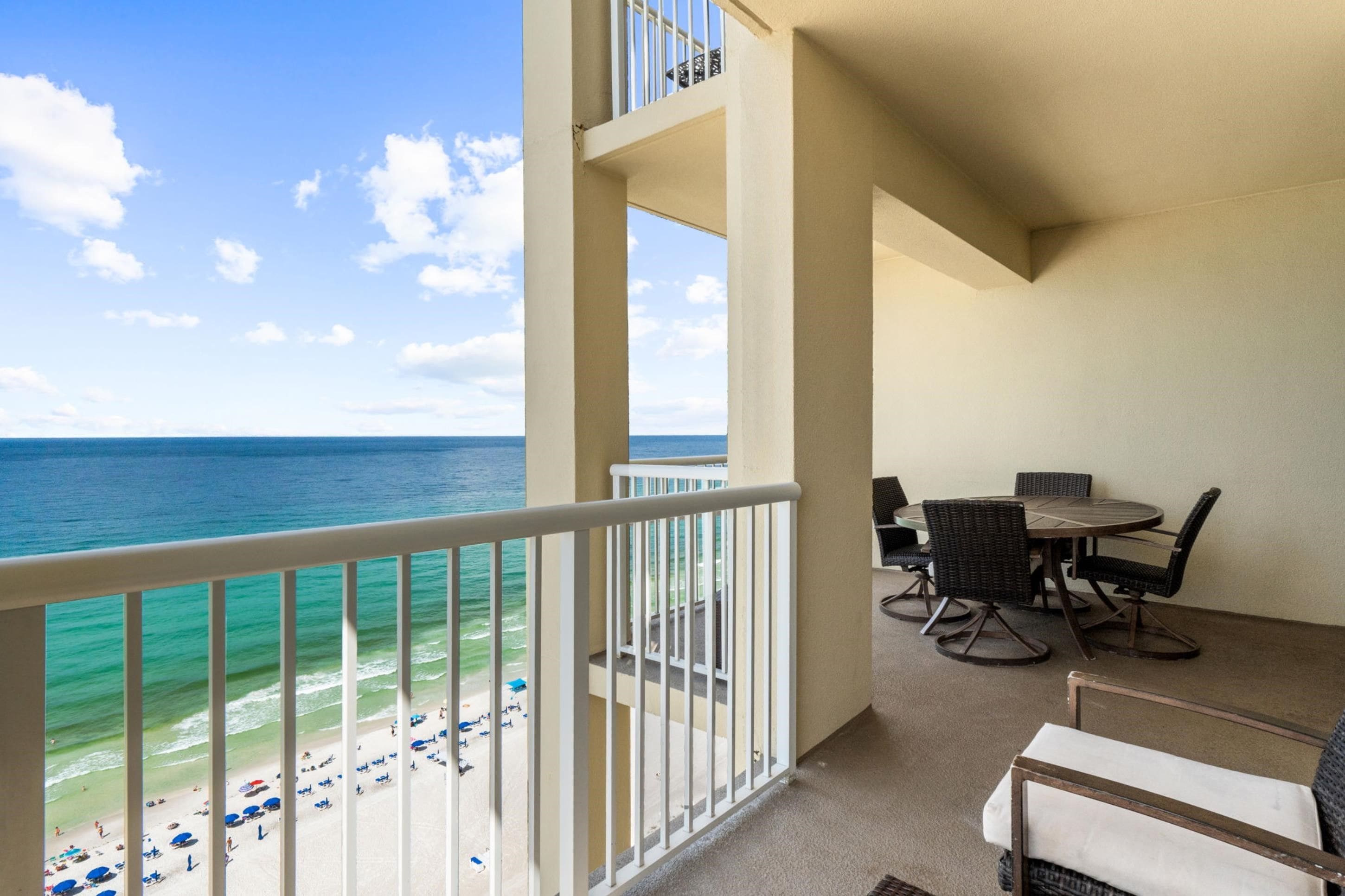 A balcony with a view of the ocean, a dining area with chairs and a table, and a beach visible in the background.