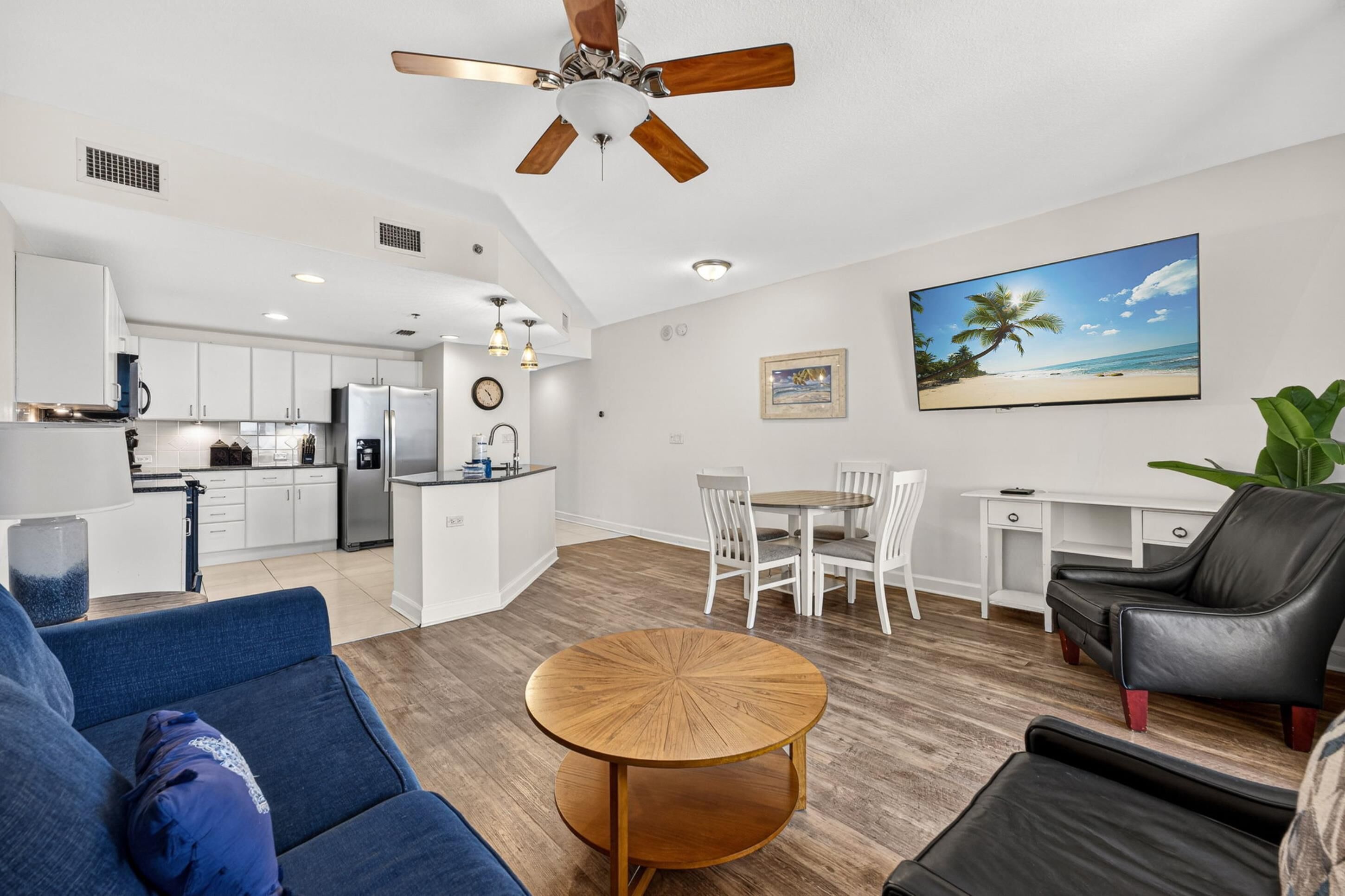 A modern kitchen with white cabinets, a dining area with a round wooden table, and a living room with a blue sofa and a black leather chair.