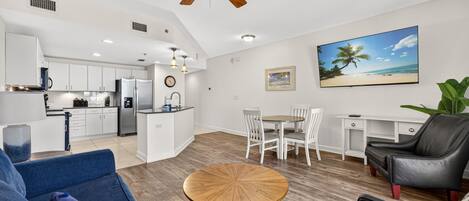 A modern kitchen with white cabinets, a dining area with a round wooden table, and a living room with a blue sofa and a black leather chair.