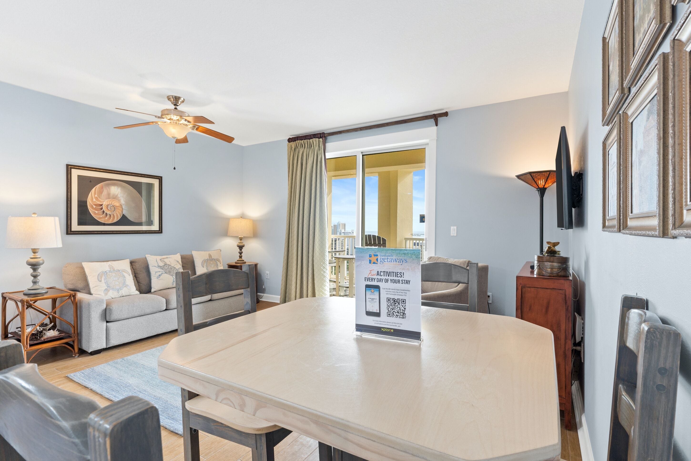 Hotelkamer met een eettafel, een bank, een plafondventilator, een lijst met een afbeelding, een lamp en een bord waarop dagelijkse activiteiten staan aangekondigd.