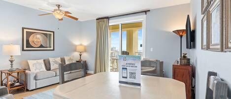 Hotelkamer met een eettafel, een bank, een plafondventilator, een lijst met een afbeelding, een lamp en een bord waarop dagelijkse activiteiten staan aangekondigd.