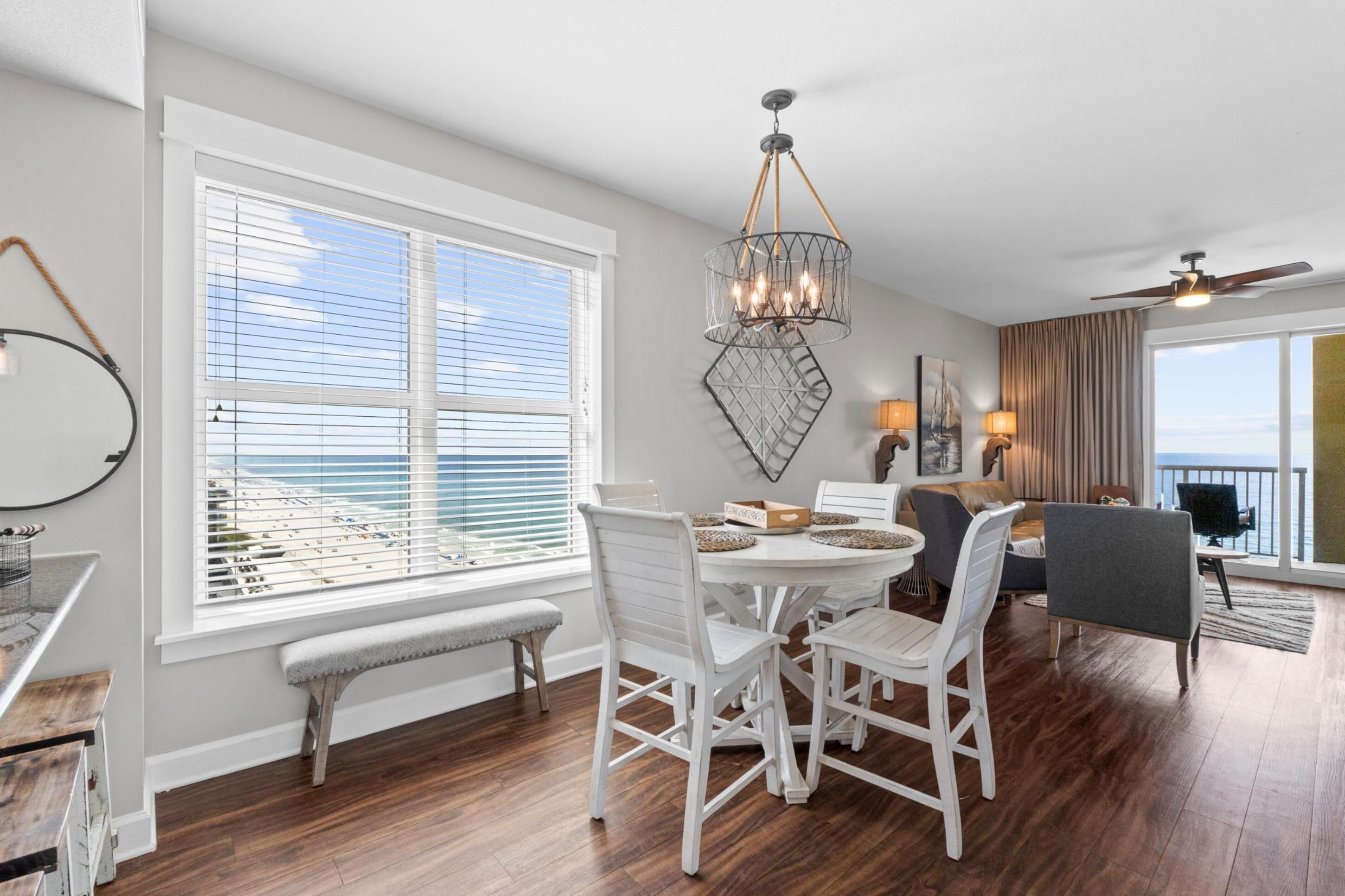 A dining area with a round table, white chairs, a chandelier, and a view of the ocean through large windows.