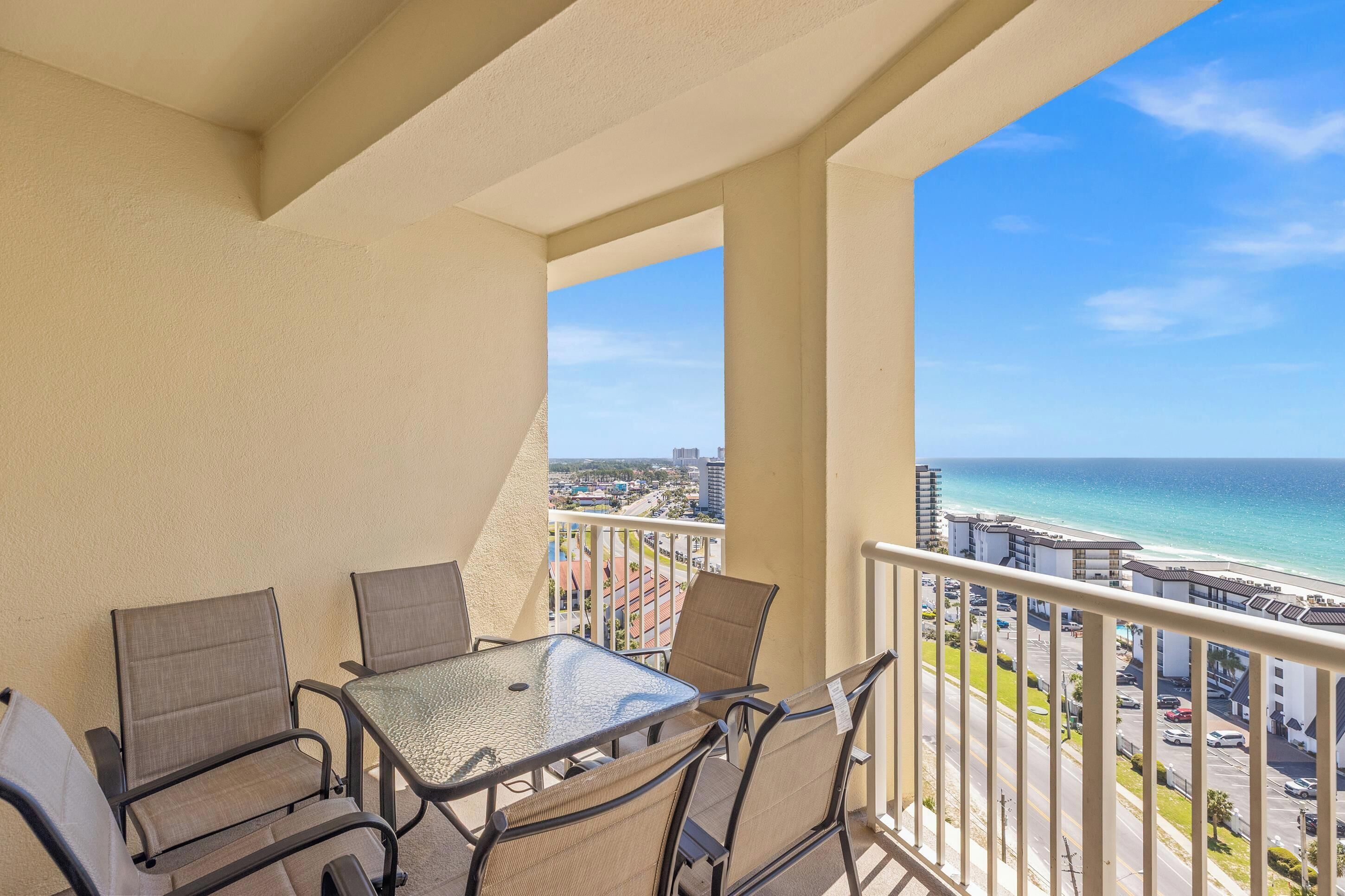 A balcony with a glass table and chairs, overlooking a beach and cityscape.