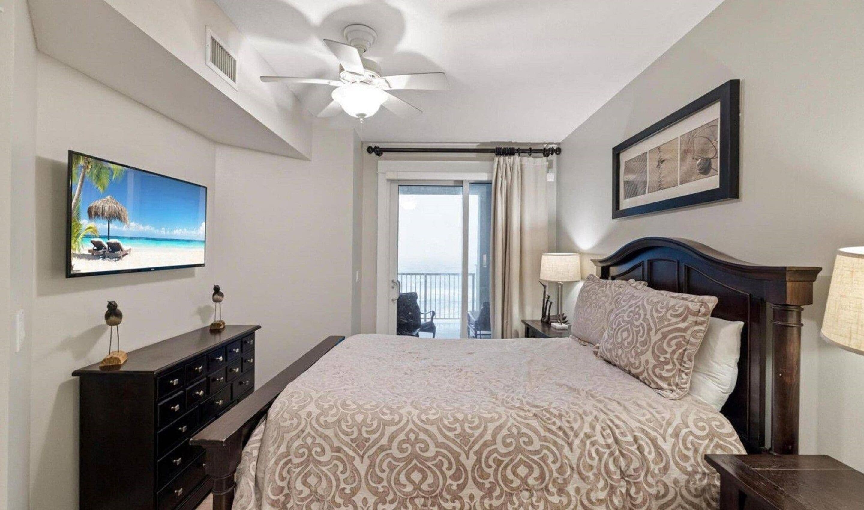 Habitación de hotel con cama, cómoda, ventilador de techo, televisor, balcón con vistas y un cuadro en la pared.