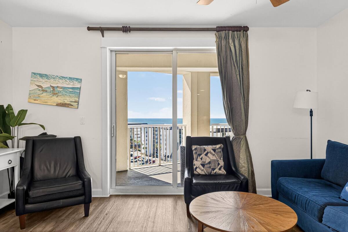 A room with a balcony, a blue sofa, a black leather chair, a wooden coffee table, and a painting of a beach scene.
